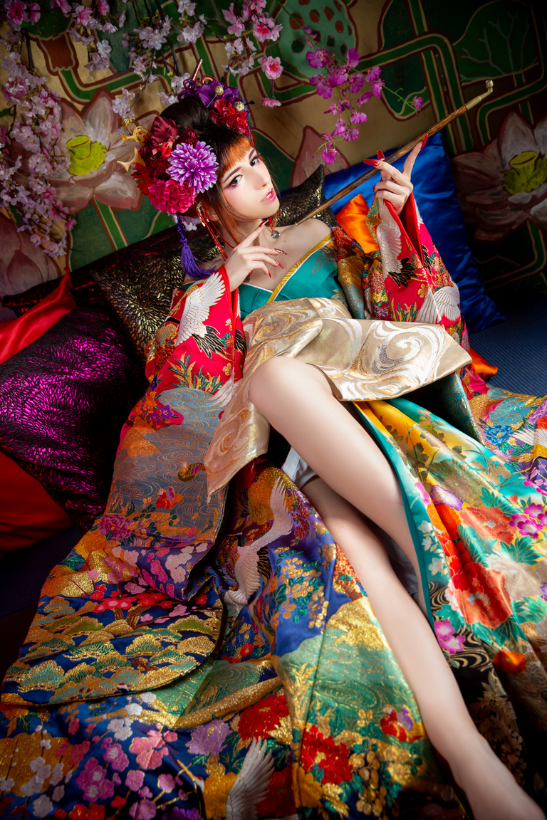 A woman dressed in a vibrant, traditional kimono adorned with intricate patterns, lounging on colorful cushions surrounded by floral decorations.