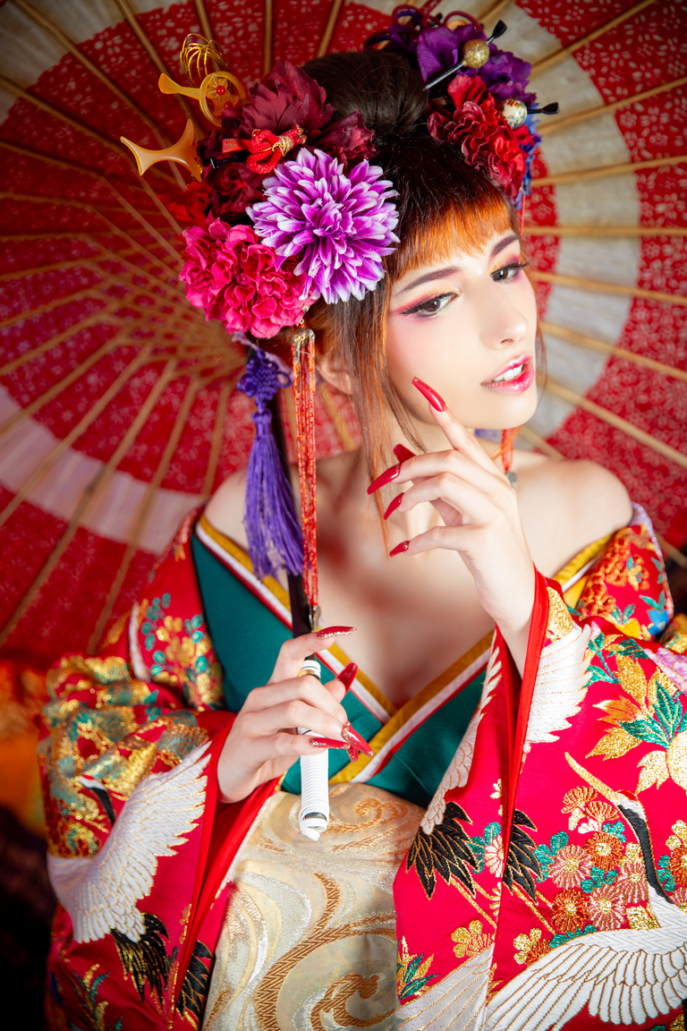 A woman dressed in a colorful kimono adorned with intricate floral patterns, holding a traditional Japanese umbrella. She has vibrant floral hair accessories and is striking a pose with her finger near her lips.
