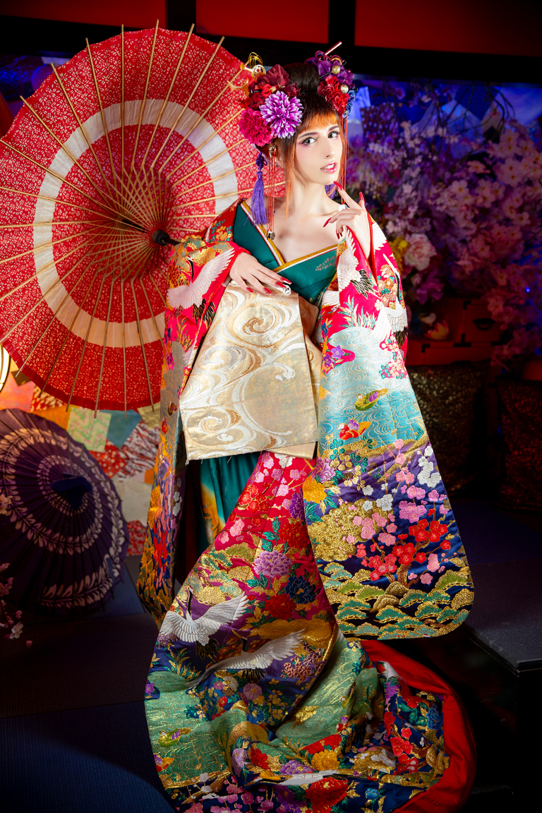 A woman in a vibrant, intricately designed kimono, holding a decorative umbrella. She has floral hair accessories and is posed against a backdrop of colorful flowers and cushions.