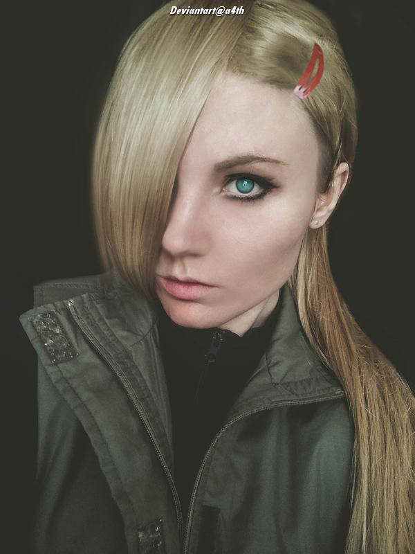 A close-up portrait of a person with long blond hair, wearing a dark jacket. They have striking blue eyes and a serious expression, with part of their hair styled over one eye.