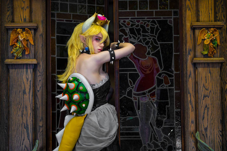 A character dressed in a costume inspired by a video game, featuring long blonde hair, a black dress, and green spiked accessories, posing in front of a decorative stained glass window.