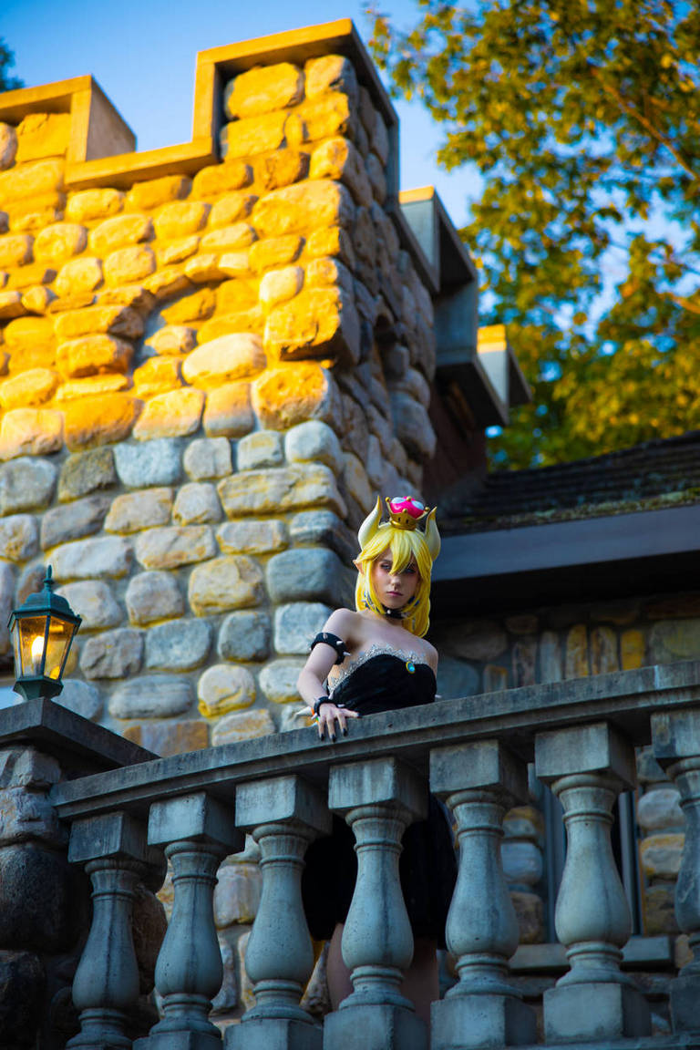 A person in a cosplay costume resembling a princess stands on a stone balcony with a castle-like structure in the background, illuminated by golden sunlight.