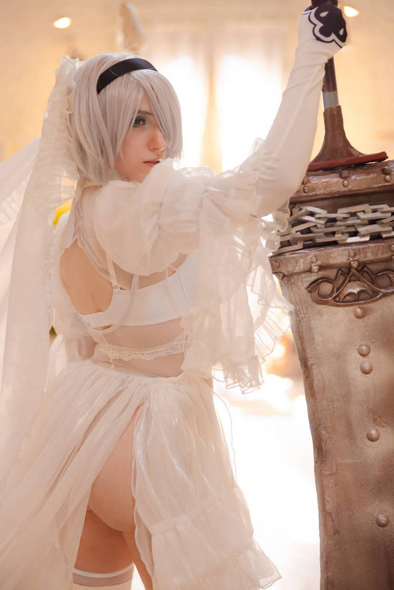 A person in a detailed costume, featuring a white dress with ruffled sleeves and a veil, holding a large weapon. The setting appears bright and soft-focused, creating an ethereal atmosphere.