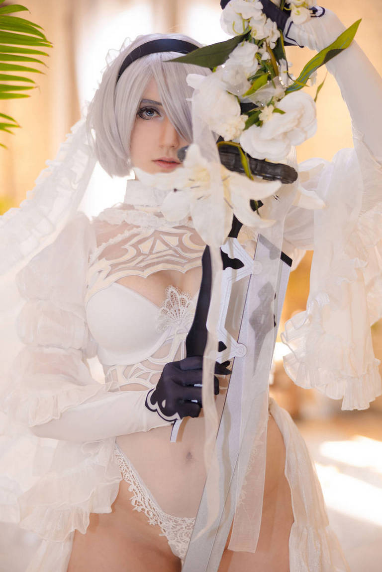 A person wearing a white and lace costume, portraying a character with short silver hair, holding a sword and surrounded by white flowers.