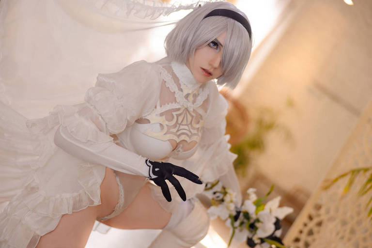 A person in a white costume with intricate designs, featuring a short gray wig and gloves, posing elegantly amidst a softly lit indoor setting.