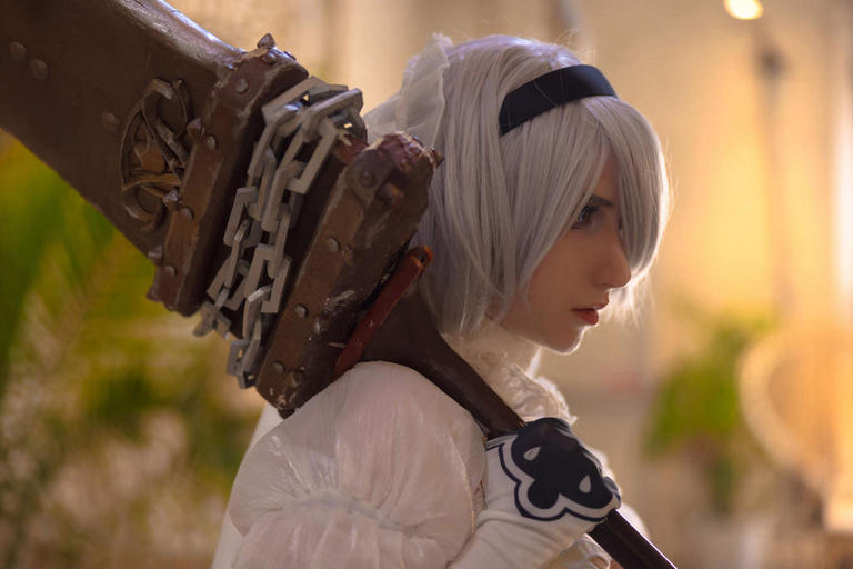 Person with white hair in a fantasy costume, holding a large weapon on their shoulder, set in a softly blurred interior.