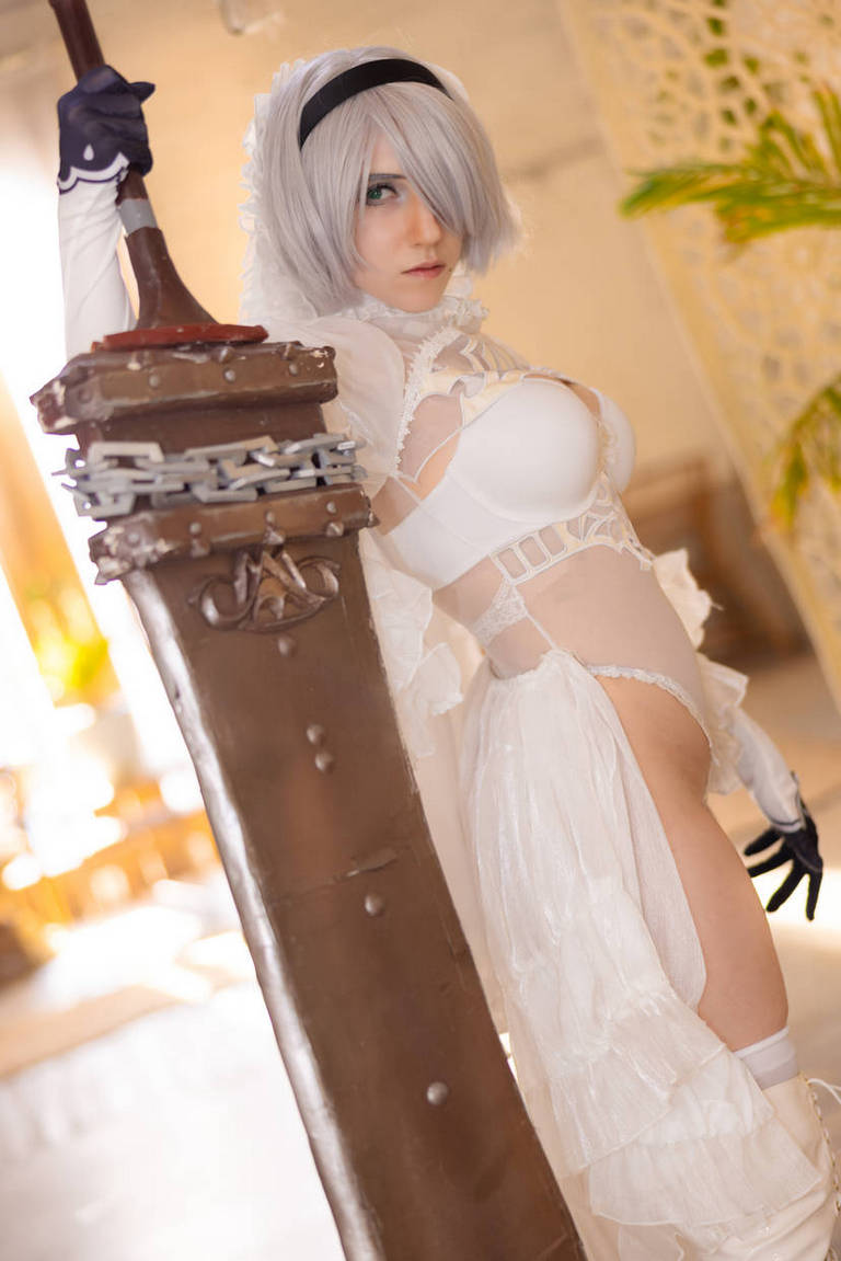 Cosplayer portraying a character with short silver hair wearing a white outfit, holding a large sword in an elegant pose.