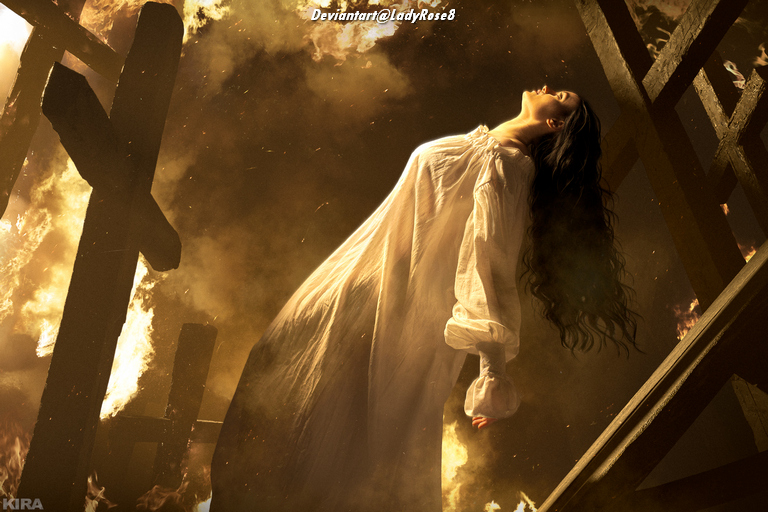 A woman in a white dress stands with her head tilted back, surrounded by flames and dark wooden structures, suggesting a dramatic and intense atmosphere.