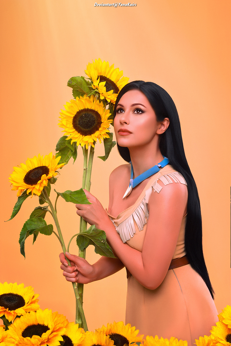 A woman in a light-colored dress with fringe holds sunflowers, gazing upward against a warm orange background.