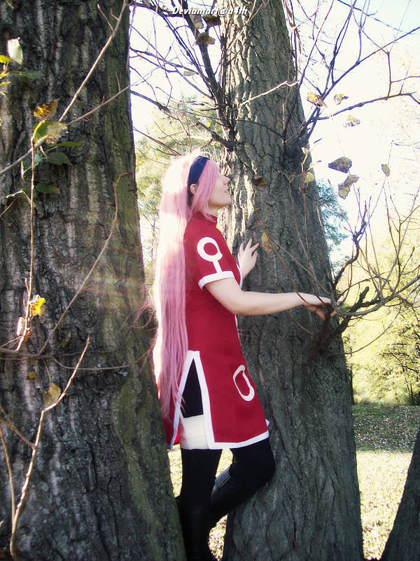 A person with long pink hair wearing a red outfit stands between two trees, reaching out to touch the trunk. The setting is outdoors with natural sunlight filtering through the foliage.