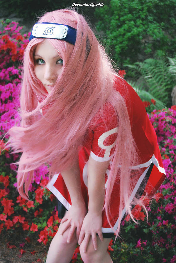 A person with long pink hair wearing a red and white outfit, posing outdoors in front of colorful flowers.