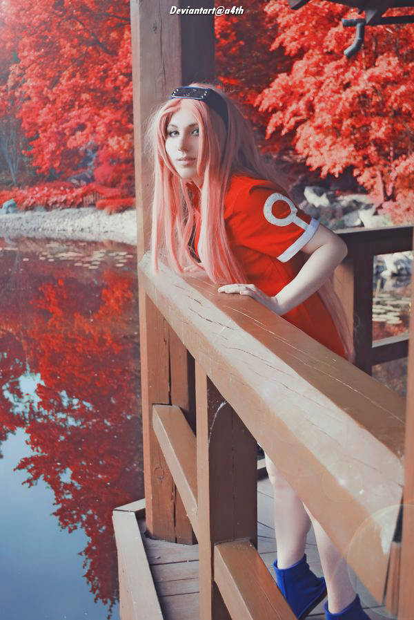 A person with long pink hair wearing a red top and blue shoes leans on a wooden railing, surrounded by vibrant red foliage and a reflective body of water.