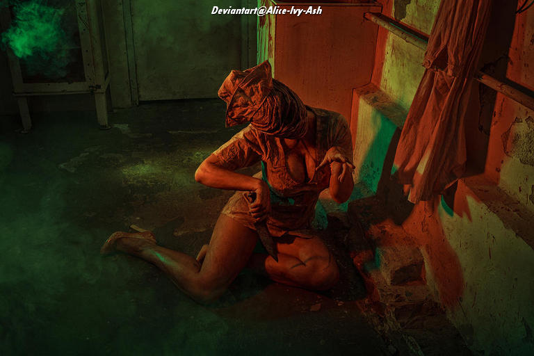 A person in a ragged outfit and a hood crouches on the floor in a dimly lit, atmospheric environment with green and orange lighting.