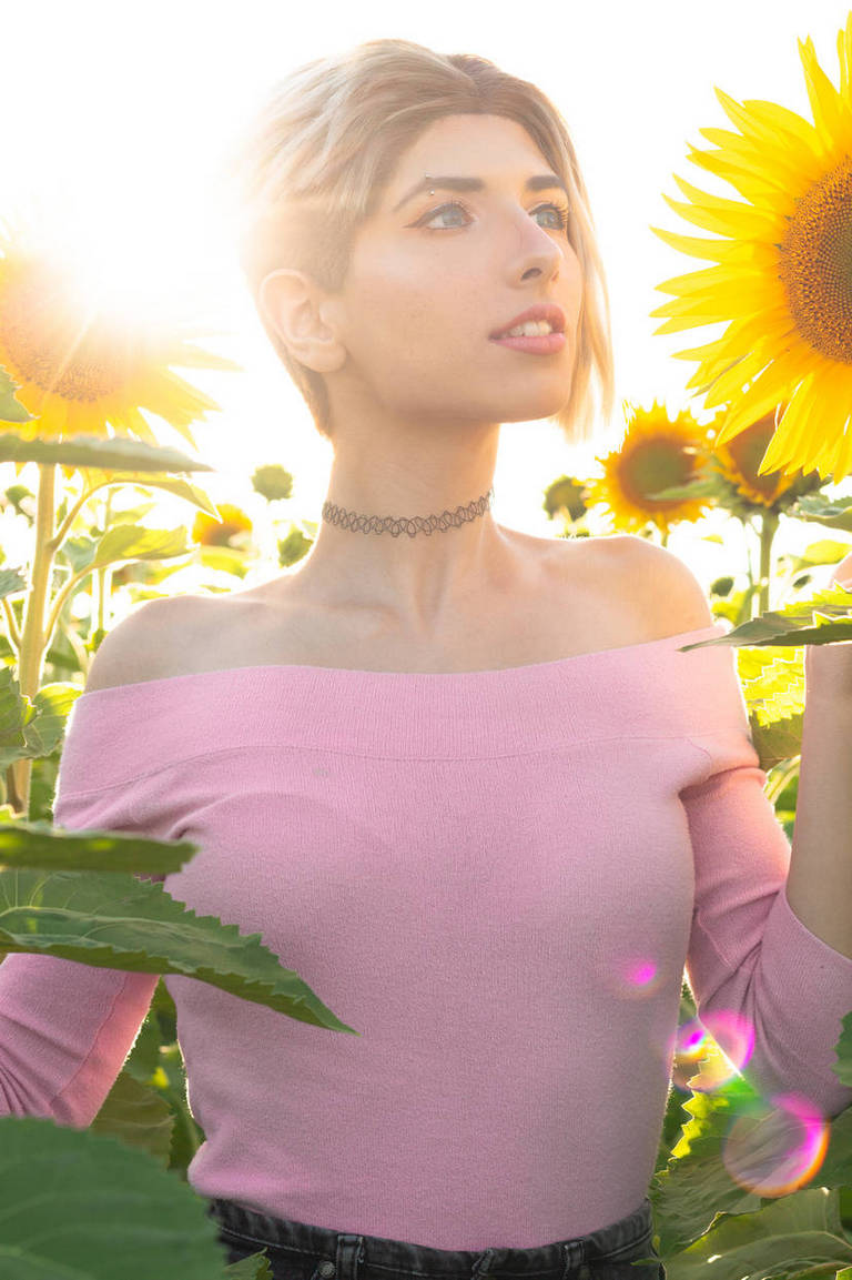 A person with short hair wearing a pink off-shoulder top stands in a sunflower field, holding a sunflower, with sunlight illuminating the scene.