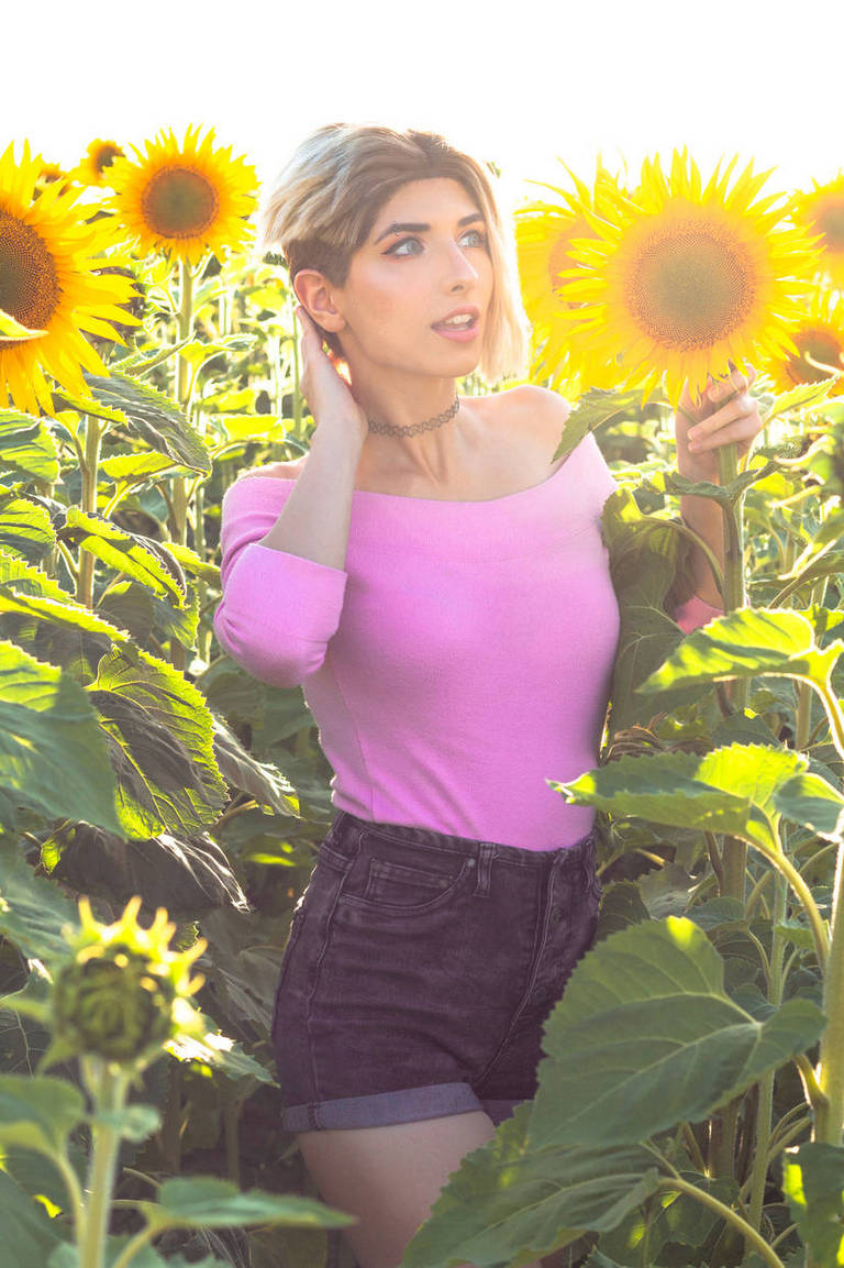 A young woman in a pink off-the-shoulder top and denim shorts, standing amidst tall sunflowers, with sunlight creating a warm glow.