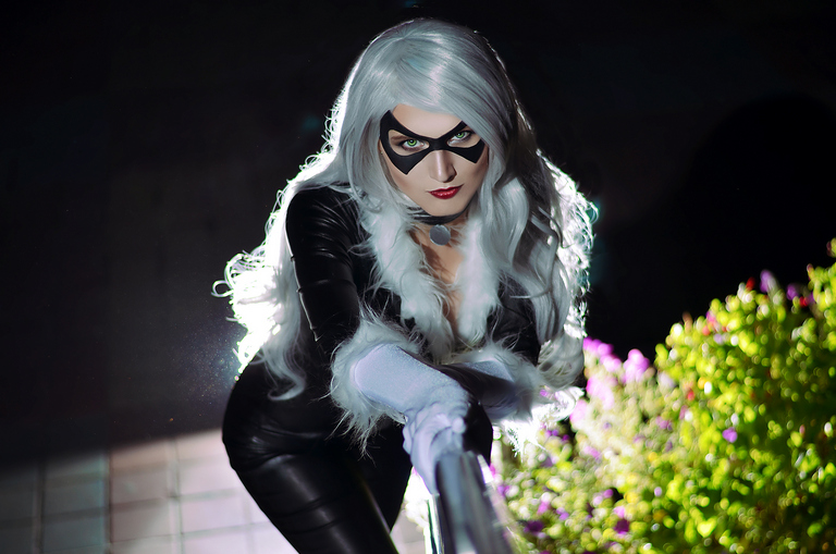 A person dressed as a character with long silver hair and a black costume, featuring a mask and white gloves, poses dramatically in a garden at night.