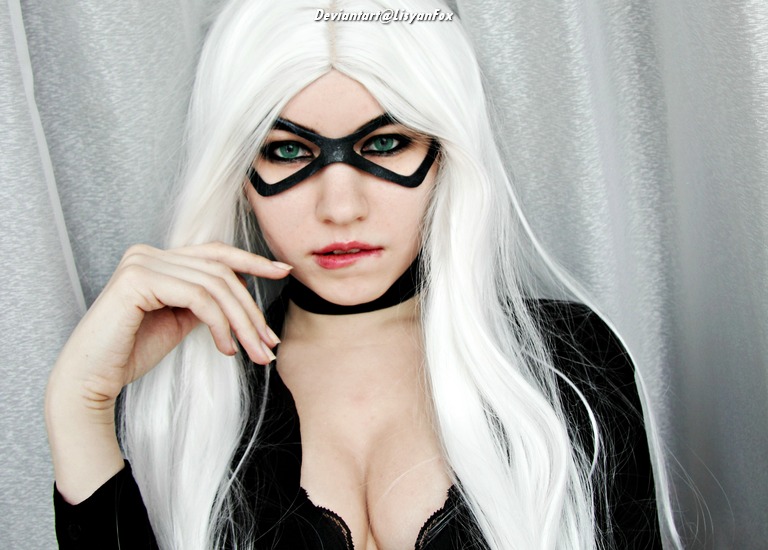 A person with long white hair and striking green eyes, wearing a black costume with a mask and choker, poses confidently against a silver backdrop.