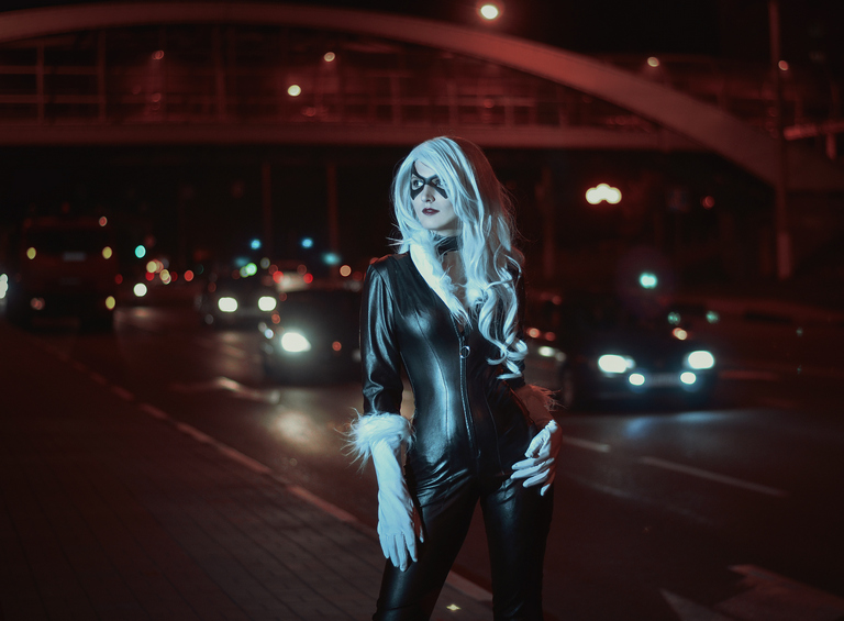 A woman in a sleek black costume with white fur accents and dramatic makeup, posing confidently on a city street at night, with cars and city lights in the background.
