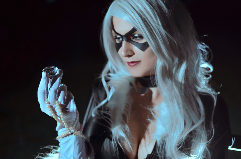 A woman with long silver hair and a black eye mask holding a shiny gemstone in her hand, dressed in a dark outfit with a pearl necklace.