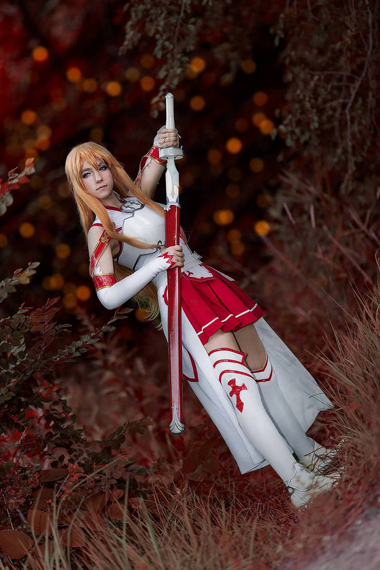 A cosplayer dressed as a character with long blonde hair, holding a sword and wearing a red and white outfit, posed in a natural setting with blurry orange lights in the background.