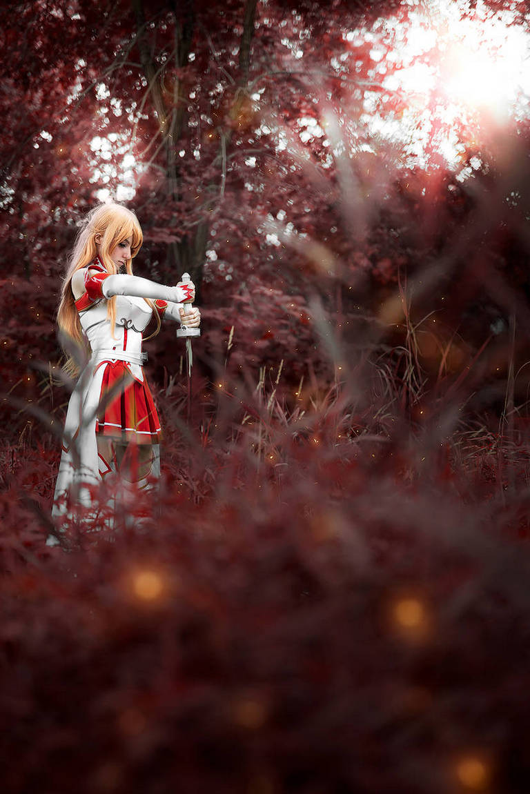 A cosplayer in a red and white outfit stands in a forest with red foliage, holding a sword against a backdrop of soft, glowing light.