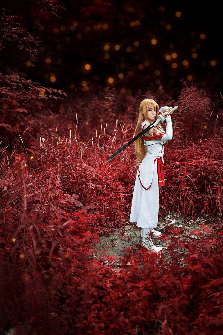 A person in a white outfit with red accents holds a sword, standing in a field of vibrant red plants against a dark, blurred background.