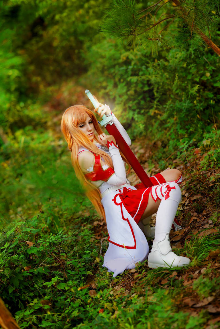 A cosplayer dressed as a character with long blonde hair and a red and white outfit, holding a sword, posed in a green forest setting.