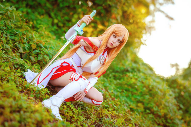 A character in a white and red costume with long blonde hair, holding a sword, posed in a natural setting with greenery and soft sunlight.