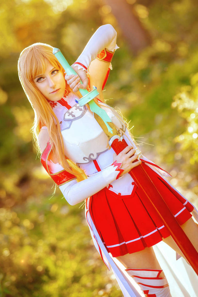 A character in a red and white outfit holding blue swords, with long blonde hair, standing in a sunlit outdoor setting.