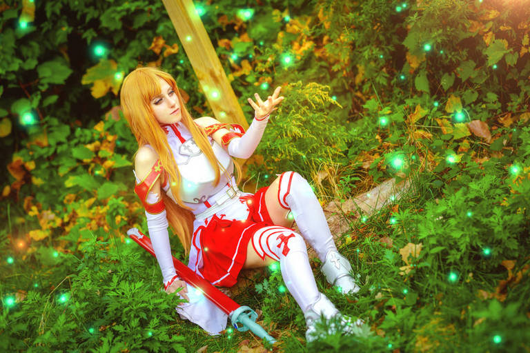 A cosplayer dressed as Asuna from Sword Art Online, sitting in a lush green setting with glowing orbs of light around her.