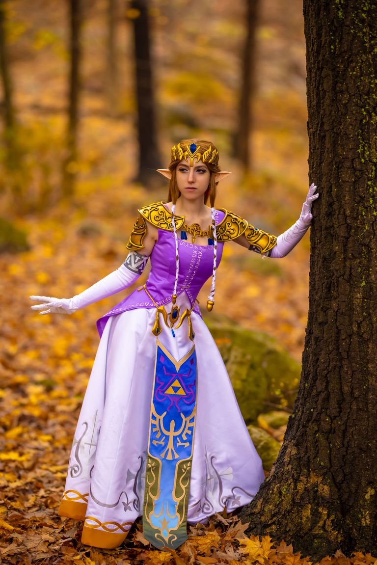 A person dressed as Princess Zelda from 'The Legend of Zelda,' posing beside a tree in a forest during autumn, wearing a purple and white costume with intricate designs.