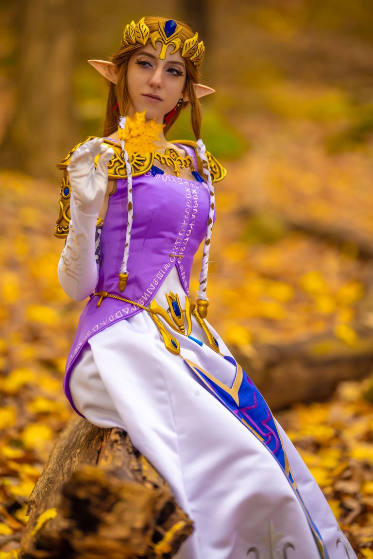 A person dressed as Princess Zelda from a fantasy game, seated on a log in a forest covered in autumn leaves, holding a yellow leaf.