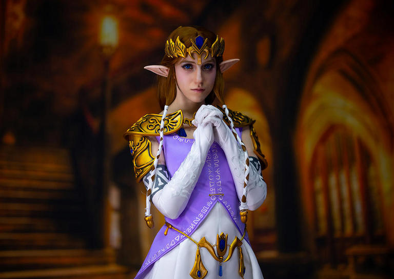 A person in elaborate fantasy costume resembling a character with long ears, golden armor and a purple gown, standing gracefully with hands clasped together against a blurred medieval-style background.