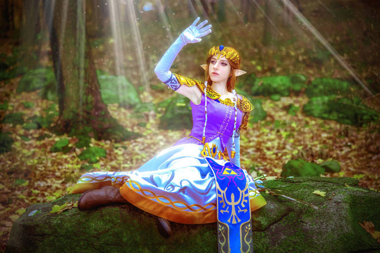 A person dressed as Princess Zelda from a fantasy game, sitting on a rock in a forest setting, with sunlight shining through the trees.