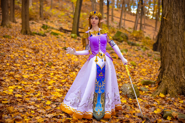 A cosplayer dressed as Princess Zelda from the video game series, standing in a forest during autumn, surrounded by falling leaves.