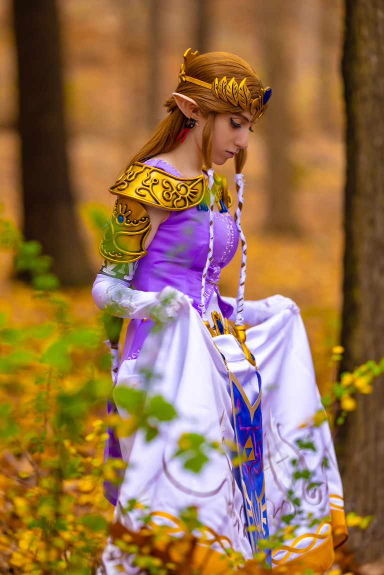 A woman dressed as Princess Zelda stands in a forest, holding her flowing white and purple gown while looking down thoughtfully.