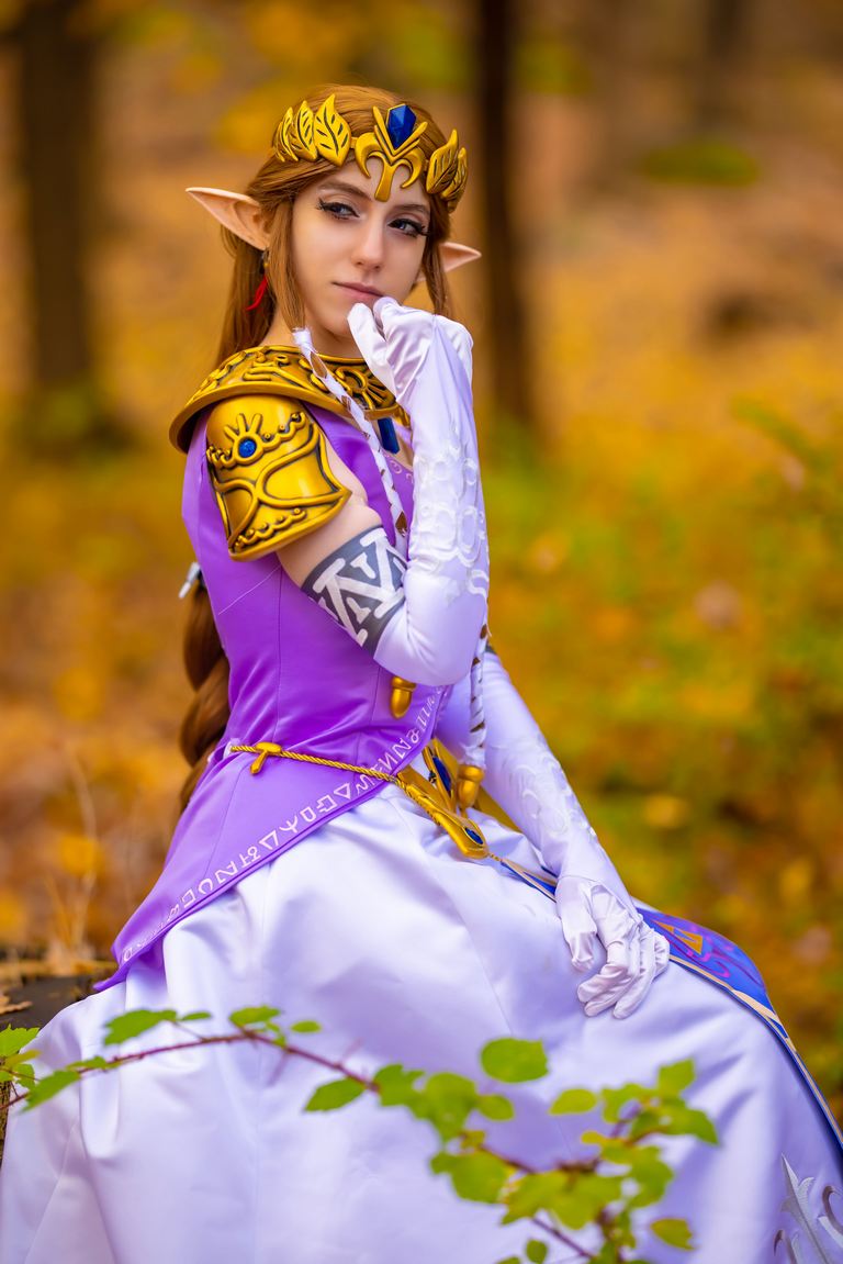 A person dressed as Princess Zelda, wearing a colorful gown with elaborate details, seated in a forest setting surrounded by autumn foliage.