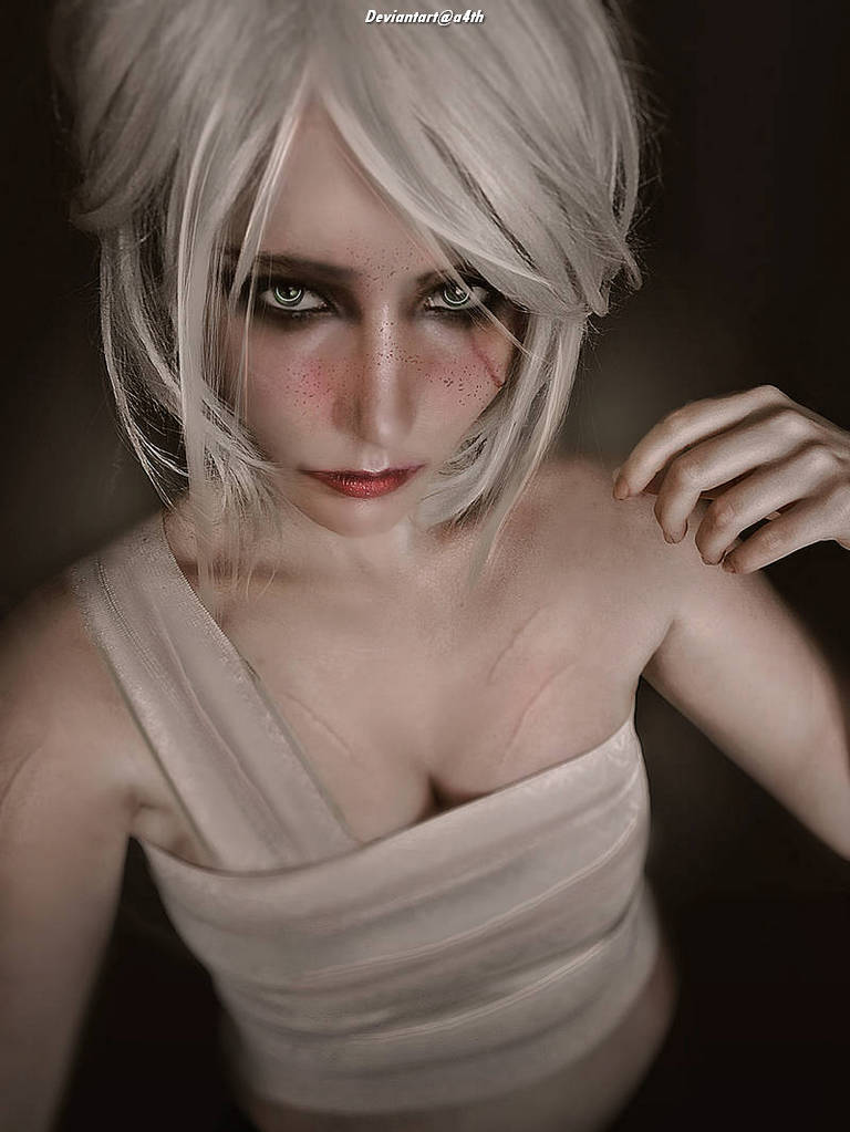 A person with short white hair and dramatic makeup, wearing a light-colored top, poses with a serious expression against a dark background.