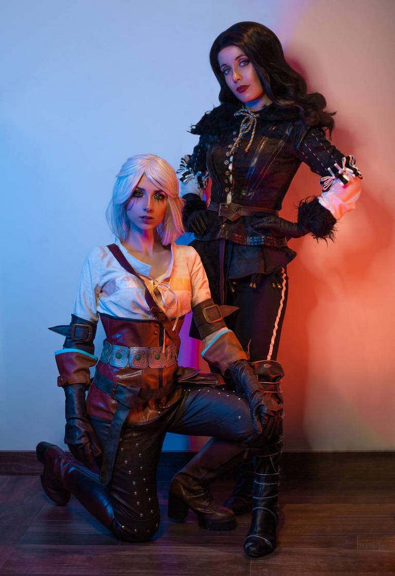 Two individuals in elaborate fantasy costumes, one kneeling with short blonde hair and the other standing with long dark hair, posing against a softly lit background with blue and pink hues.