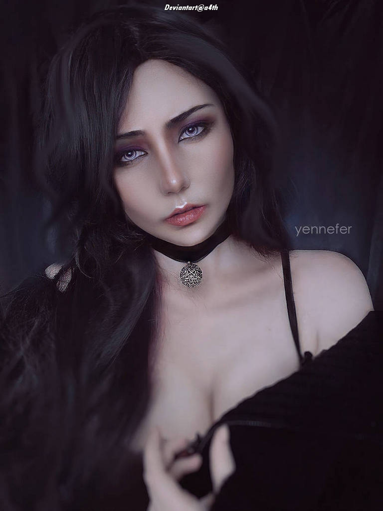 A close-up portrait of a woman with long dark hair, wearing a black off-shoulder garment and a choker necklace. She has striking makeup with dark eye shadow and a subtle lip color, set against a dark background.