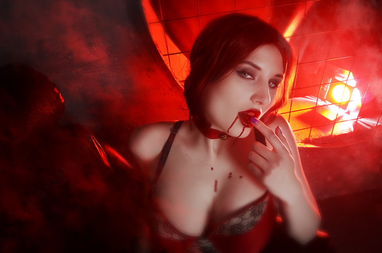 A woman with dark hair in a red setting, playfully posing with her finger in her mouth and a hint of blood on her lips, wearing a revealing outfit.