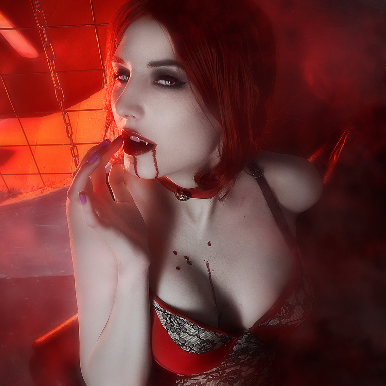 A woman with red hair and dark makeup poses dramatically, wearing a black lace outfit and a collar. There are droplets of red on her lips and chin, with a misty, dark background that enhances the moody atmosphere.
