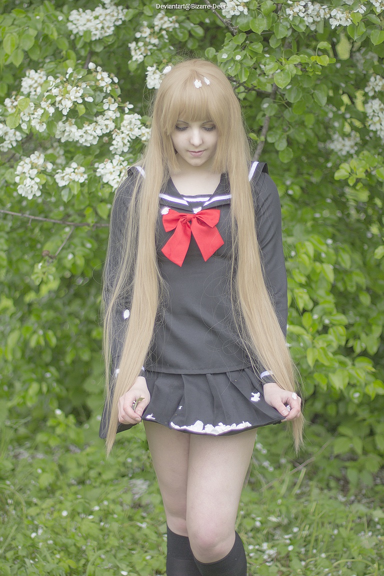 A young woman wearing a black school uniform with a red bow, standing in front of green foliage and white flowers, looking down.