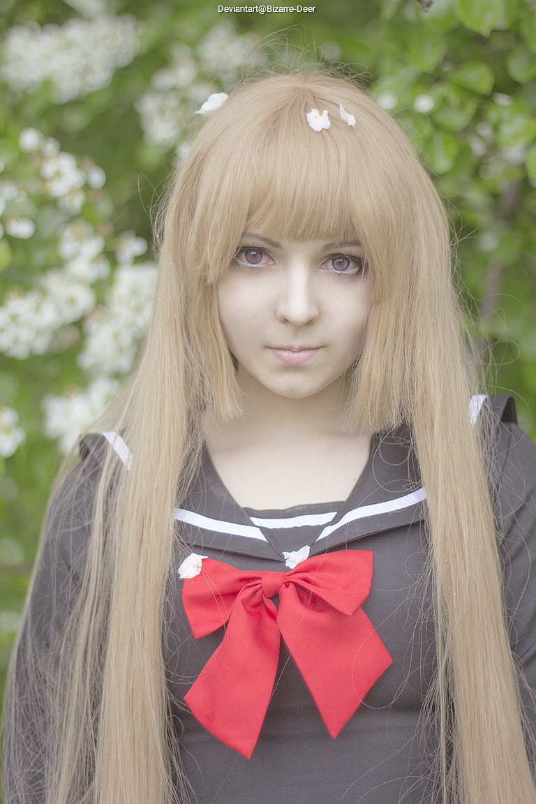 A close-up portrait of a young person with long blonde hair and a red bow, wearing a dark sailor-style outfit. The background features green leaves and white flowers.