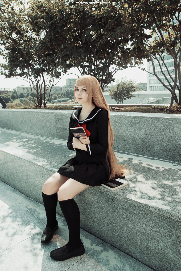 A young person in a black school uniform and long blonde hair sits on a stone bench, holding books, with trees and buildings in the background.