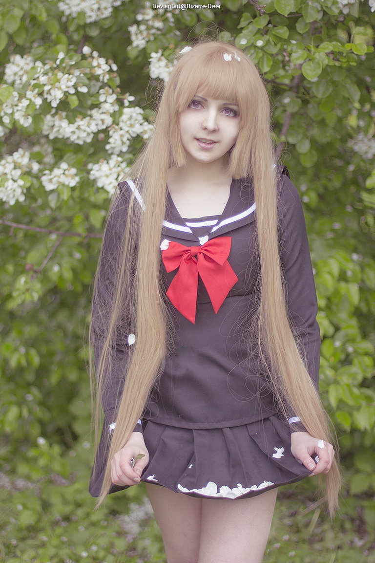 A person with long blonde hair and a black school uniform with a red bow stands in front of blooming greenery.