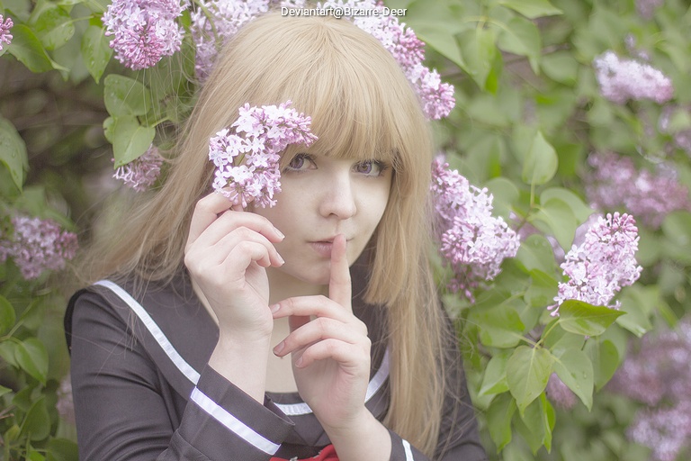 A person with long blonde hair in a black and white outfit holds lilac flowers close to their face, making a shushing gesture among a backdrop of blooming lilac bushes.