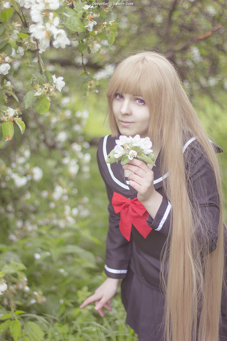 Young person with long blonde hair wearing a black dress with a red bow, holding white flowers while standing in a garden with apple blossoms.