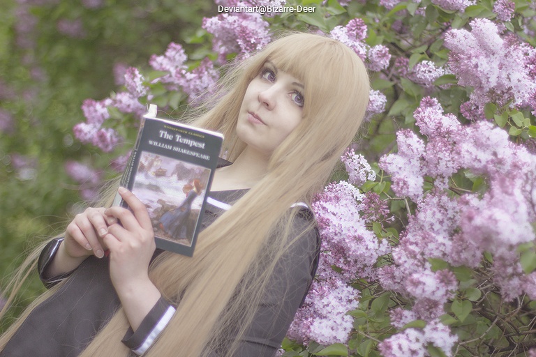 A person with long blonde hair holding a book titled 'The Tempest' by William Shakespeare, standing in front of blooming lilac flowers.