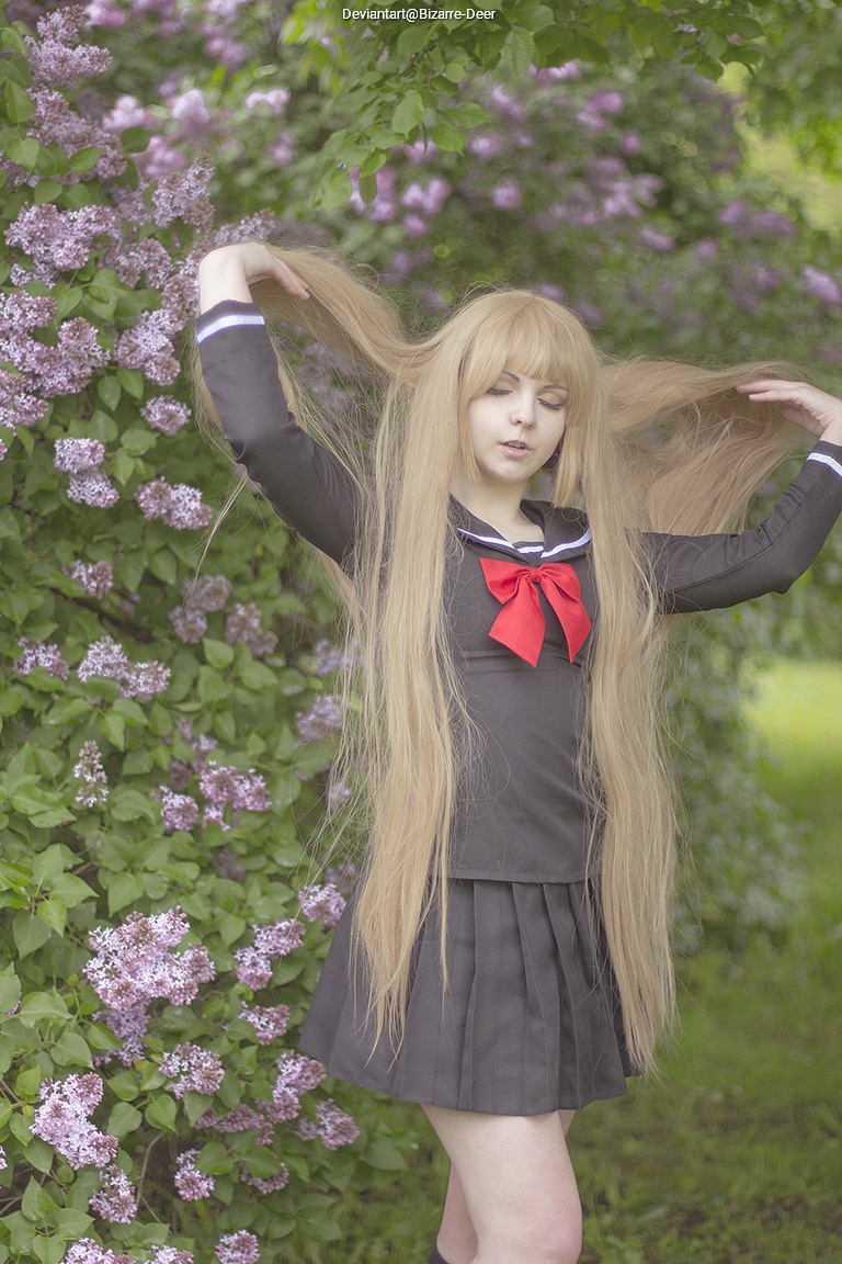 A person in a school uniform with long blonde hair posing outdoors in front of flowering lilac bushes.